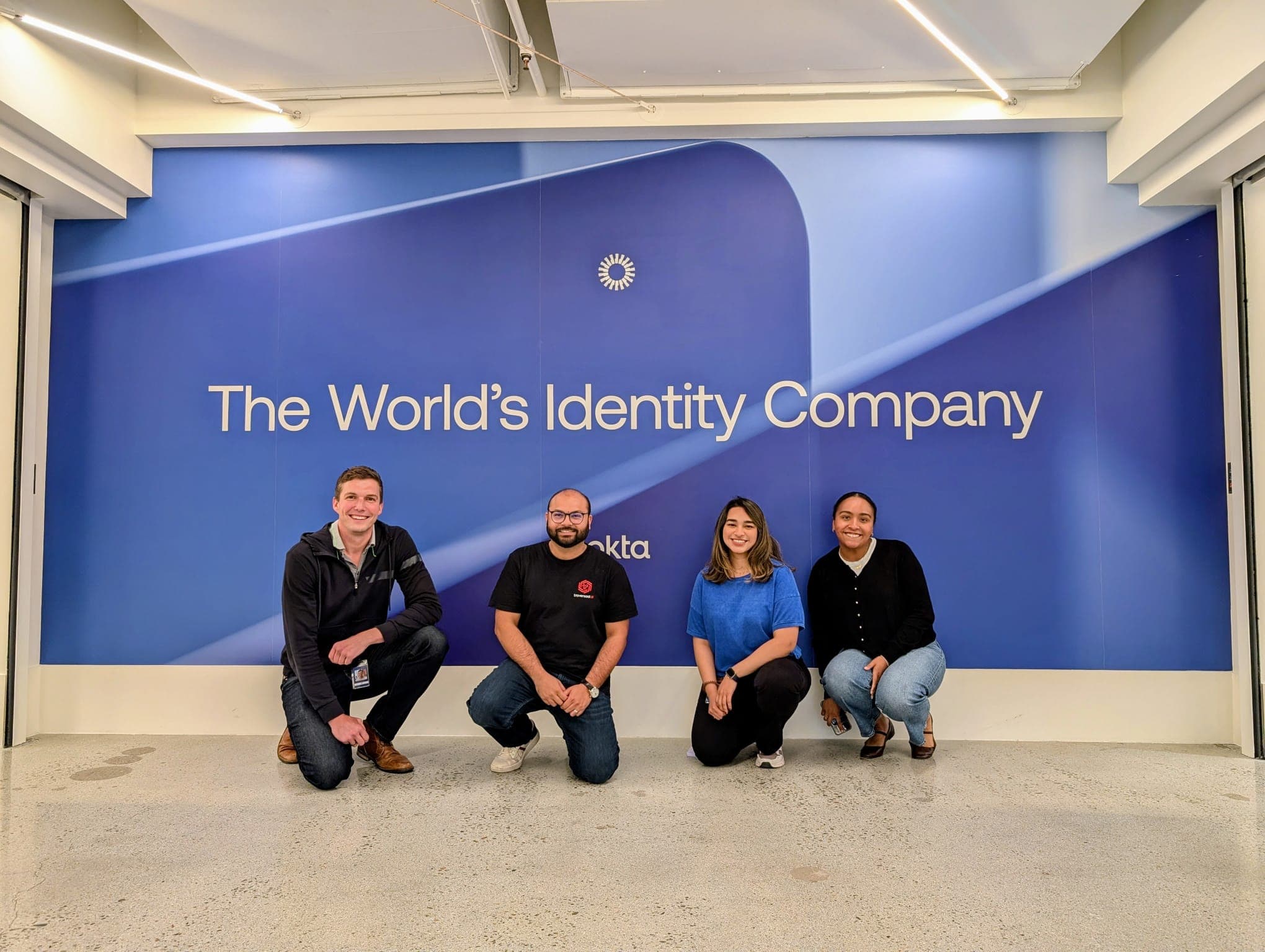 Hamza Farooq with three members of the Okta team in front of the Okta wall